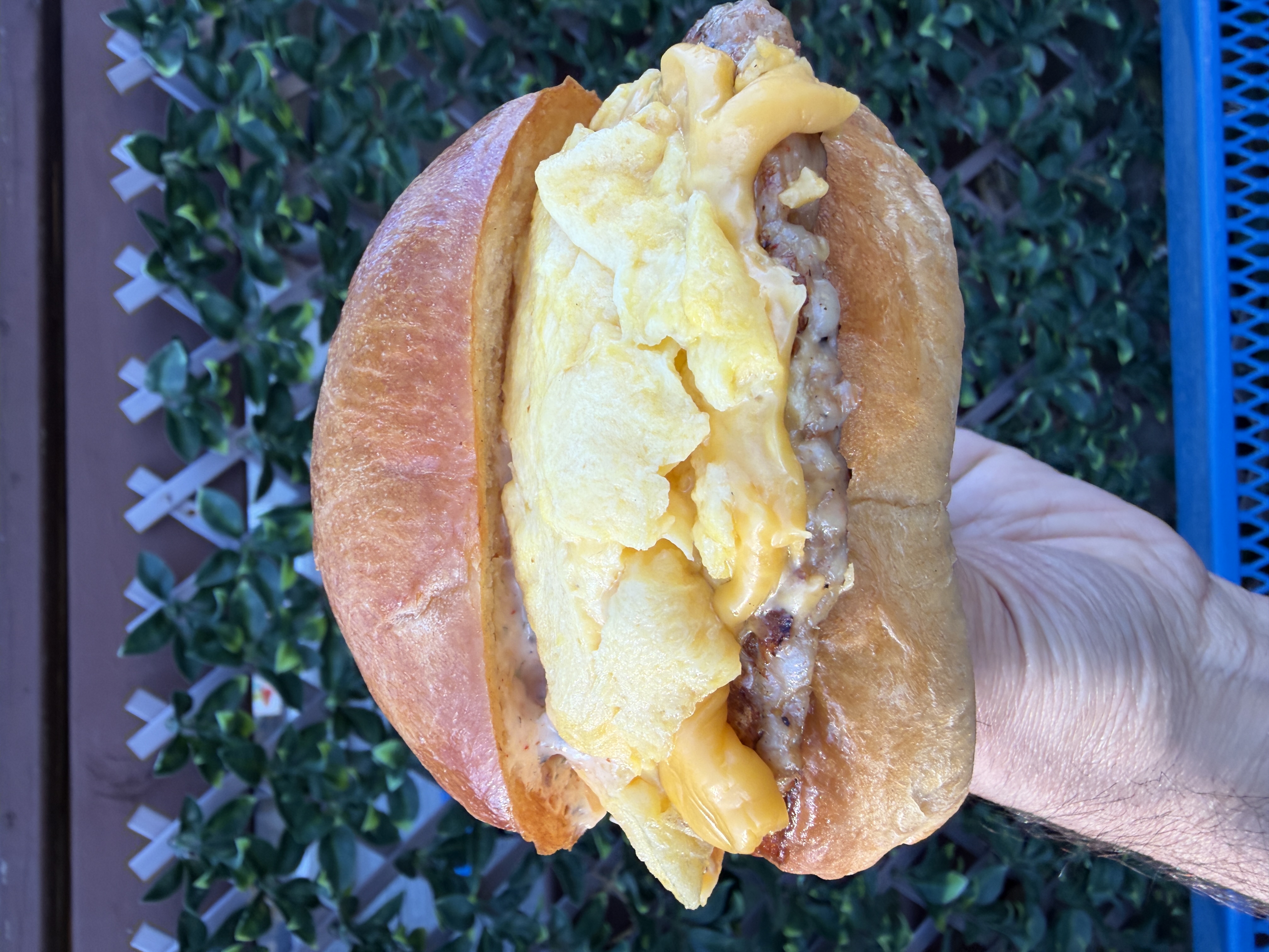 A close-up shot of a hand holding a very large breakfast sandwich containing a sausage patty, abundant scrambled eggs and melted American cheese.