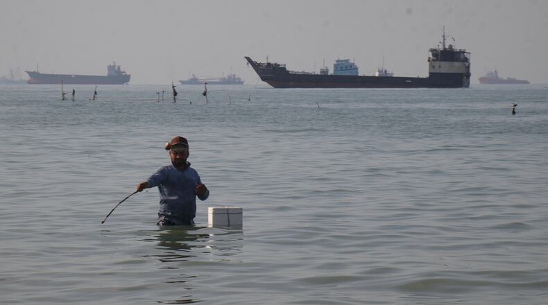 A man stands in the water, appearing to fish, as bulk carriers, cargo ships, and service vessels line the horizon in the Strait of Hormuz off Bandar Abbas, Iran, Monday, April 27, 2026.(Razieh Poudat/ISNA via AP)