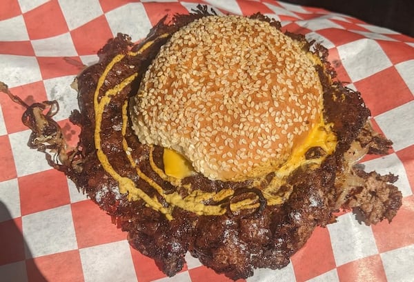 A very large smash burger that comes out about an inch beyond the edges of its sesame seed bun is shown on a checkerboard paper. It has cheese and a spiral of mustard peeking out from the top bun.