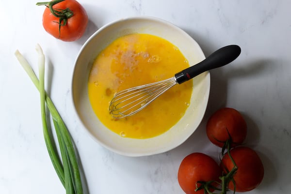 A whisk is balanced over a bowl holding beaten eggs. Four whole tomatoes and two green onions are artfully arranged around the bowl.