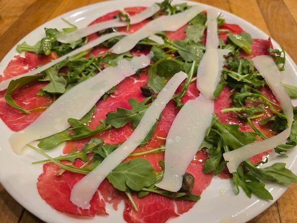 A plate of beef carpaccio with thin slices of beef topped with strips of parmesan, capers and arugula.