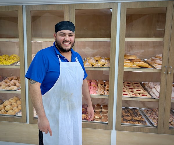 A baker in an apron and hat stands in front of cases of baked goods