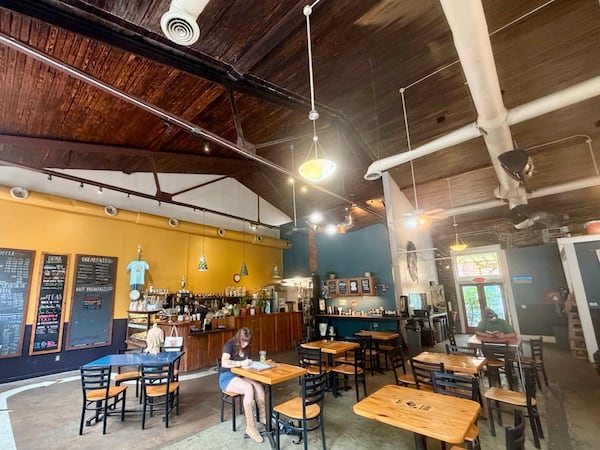 The interior of a coffee shop called the Sentient Bean in Savannah, with high ceilings, wood-topped cafe tables and a chalkboard menu next to the coffee bar.