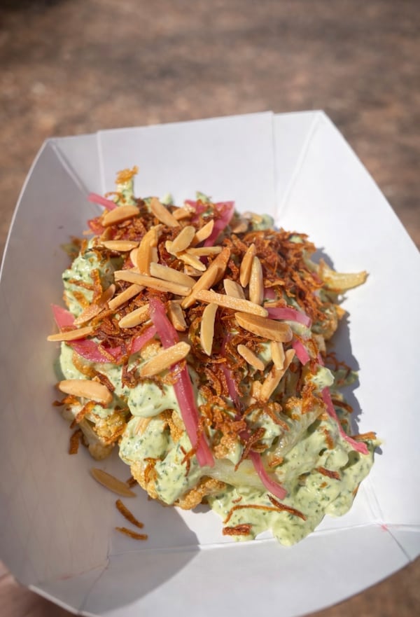 A paper food boat holds a cauliflower-herb yogurt side dish resembling potato salad. It's garnished with large ieces of slivered almonds, fried onions and pickled onions.