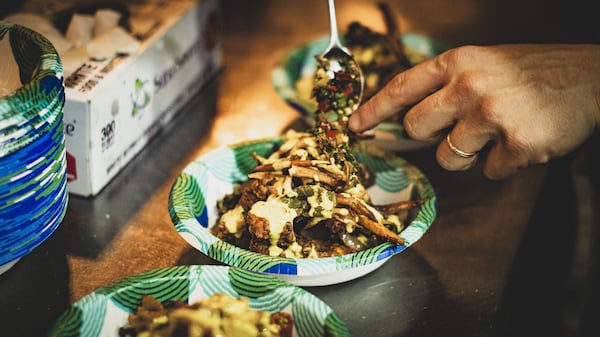 Two hands add a spoonful of garnish to a "spice bag," which is a bowl of fried chicken,  french fries, vegetables and sauce.
