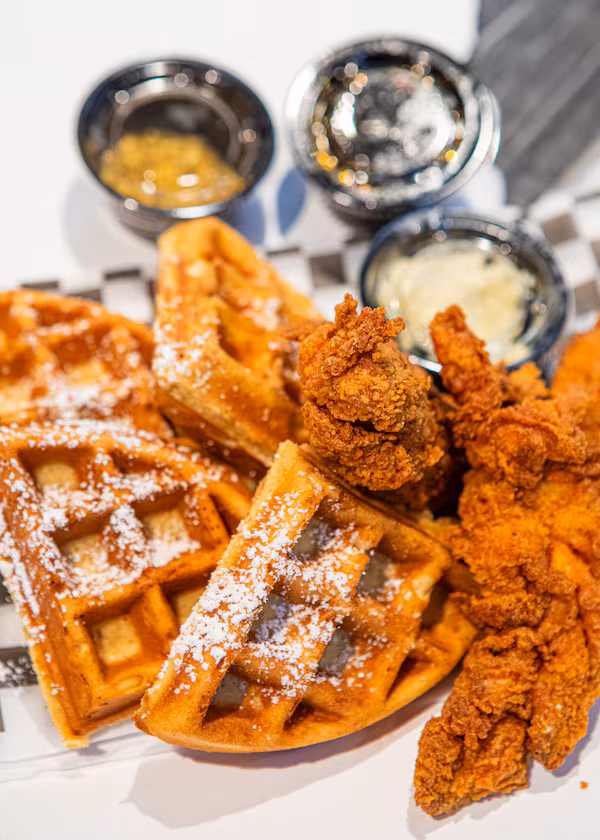 A close-up shot of waffle quarters dusted with powdered sugar, and fried chicken pieces next to the waffles. Plastic containers of butter and syrup are in the background.