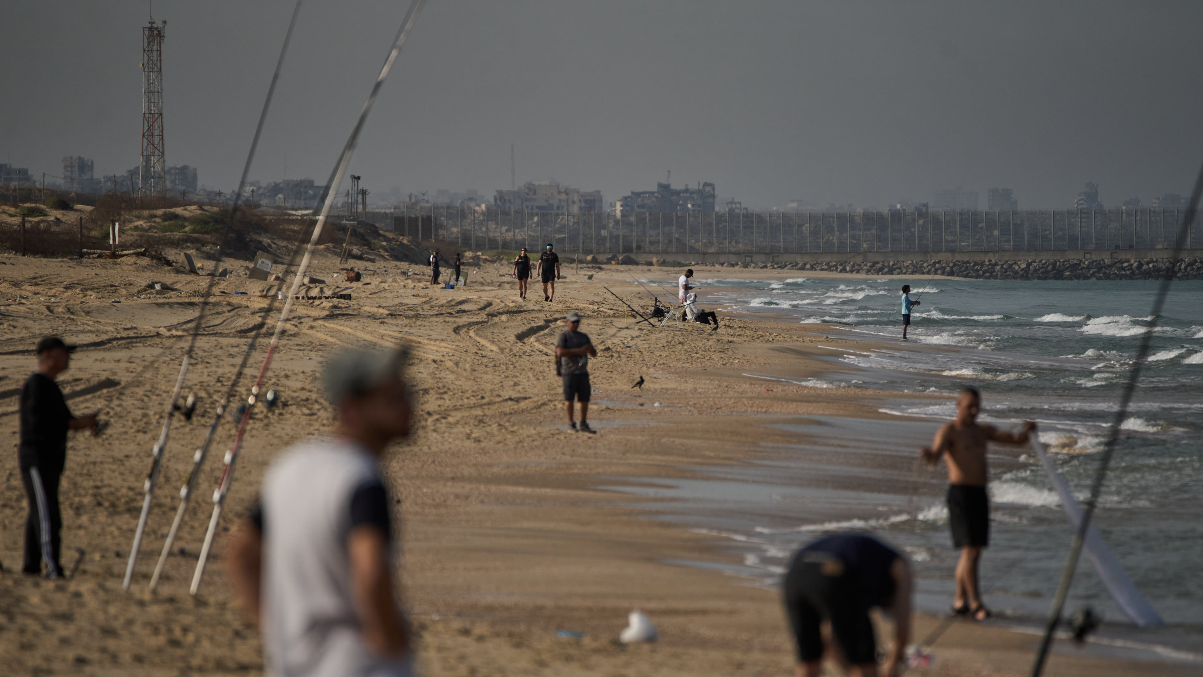 Backdropped by buildings destroyed during Israeli ground and air operations in the northern Gaza Strip, people walk and fish at Zikim Beach, near Ashkelon, in southern Israel, Friday, Oct. 17, 2025. (AP Photo/Leo Correa)