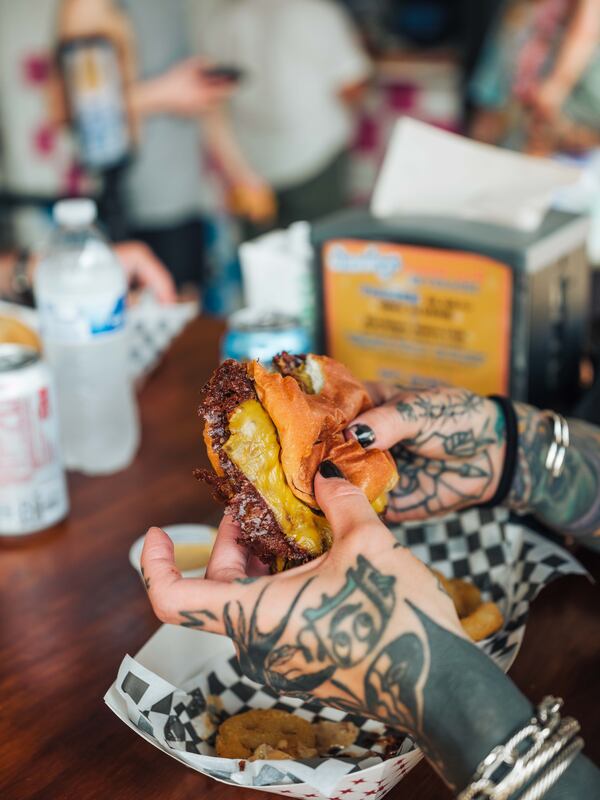 A pair of tattooed hands hold a smashburger in a restaurant.