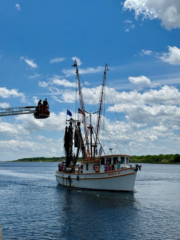 Two people on a crane in the upper left corner of the photo look over a shrimp boat in the water beneath them.