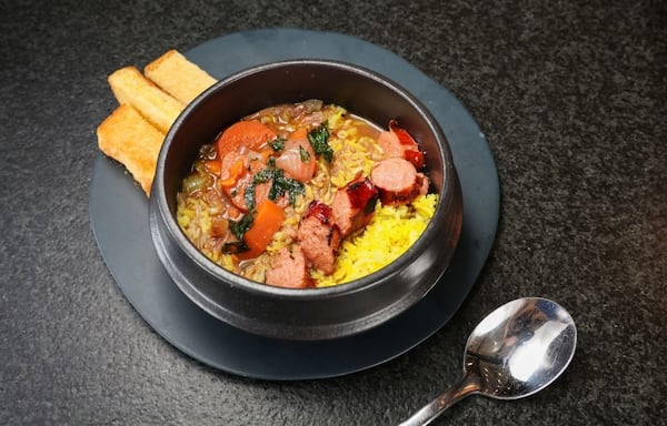 A bowl of gumbo served with toast and a spoon. Grilled sausage features prominently in the gumbo, as well as yellow rice and vegetables.