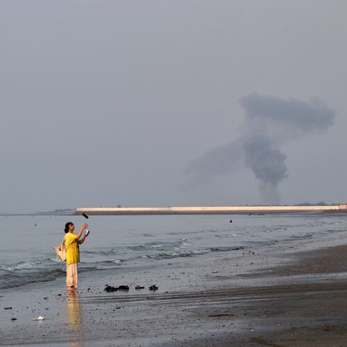 A woman stands at the water's edge along the Strait of Hormuz as a plume of smoke rises in the background following an explosion, off Bandar Abbas, Iran, Monday, July 13, 2026. (Razieh Poudat/ISNA via AP)