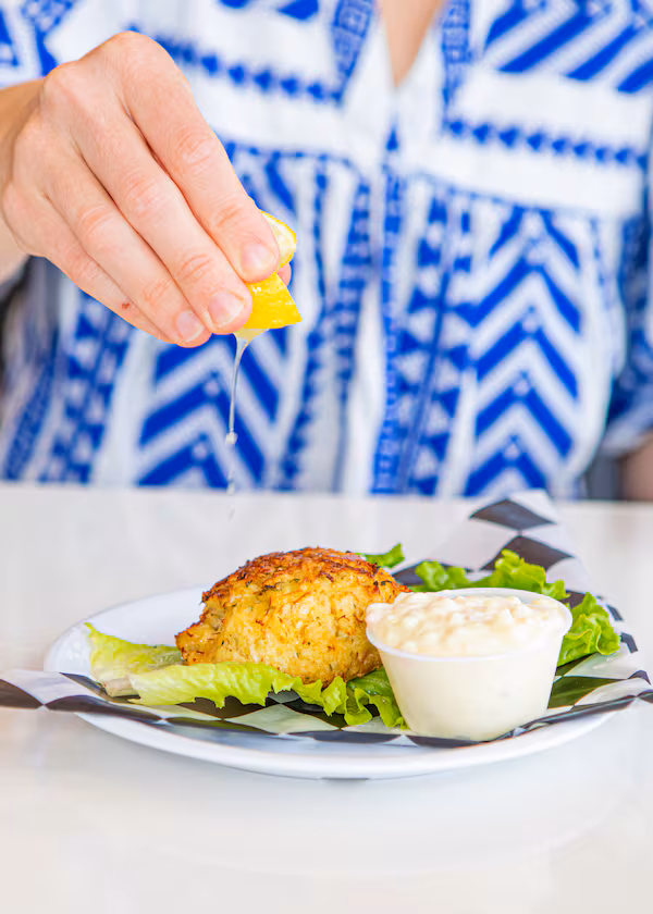 A hand squeezes a lemon wedge over a crab cake, which is plated on a lettuce leaf and served with a sauce.