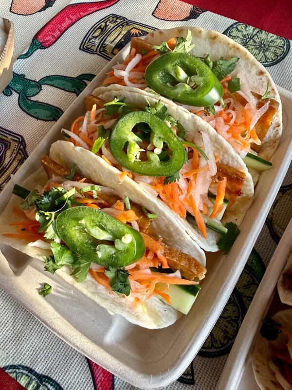 Three banh mi tacos from Karly's Kitchen are lined up in a takeaway container on a festive placemat. They are garnished with jalapeno slices.