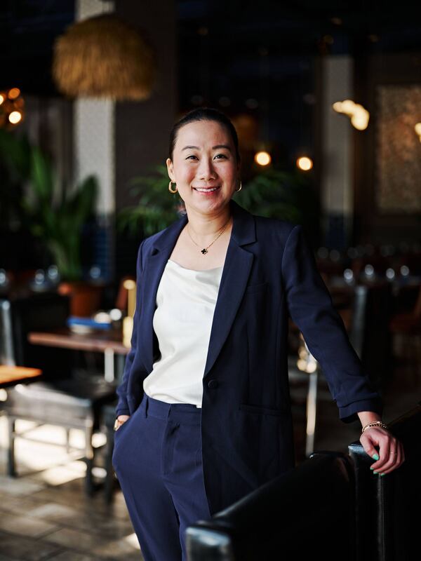 Photo of Niki Pattharakositkul dressed in a smart pants suit and tasteful gold jewelry, smiling pleasantly and standing in one of her restaurants.