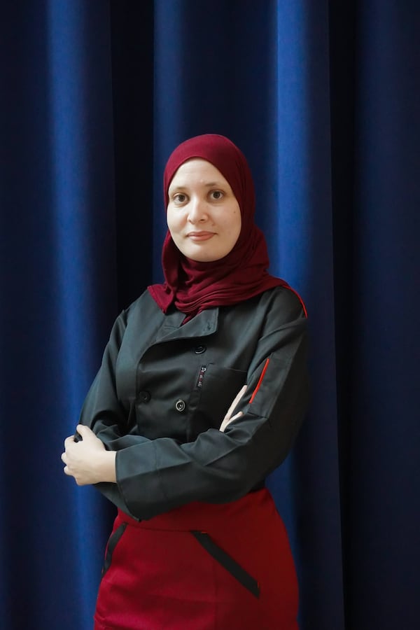 A woman in a chef's coat and hijab stands with her arms crossed looking into the camera.