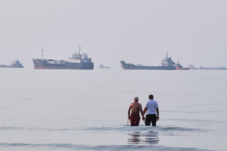 Two men wade in the waters of the Strait of Hormuz with vessels anchored in the background, off Bandar Abbas, Iran, Sunday, July 12, 2026. (Razieh Poudat/ISNA via AP)