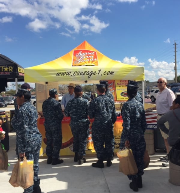 Airmen crowd around a Sunset Farm Foods pop-up tent on a bright, sunny day.