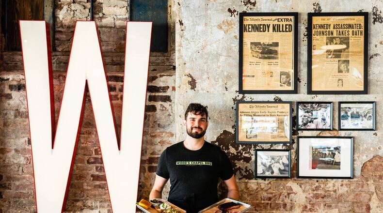 Server Zac Bodenhamer holds platters of barbecue and side dishes in front of a wall of political memorabilia at Wood’s Chapel BBQ. CONTRIBUTED BY HENRI HOLLIS