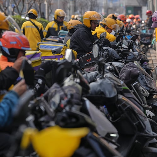 A cleaner sweeps near food delivery riders gather outside restaurants waiting for online orders, in Beijing, China, Wednesday, March 25, 2026. (AP Photo/Andy Wong)
