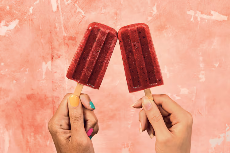 two hands holding one red popsicle each with a pink background
