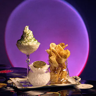 A coupe glass is filled with a whipped, creamy spread next to an open cannister of caviar on ice and a glass jar piled high with potato chips. All three vessels sit on a silver tray with a pink prismatic spotlight projected on the wall behind.