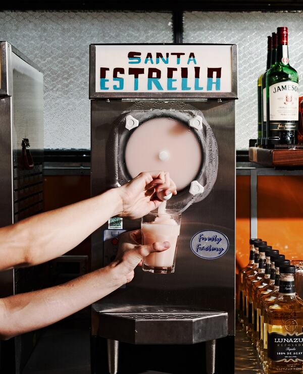Two arms extend to pour a frozen margarita into a glass out of a margarita machine. The machine has a large sign on it that says "Santa Estrella."
