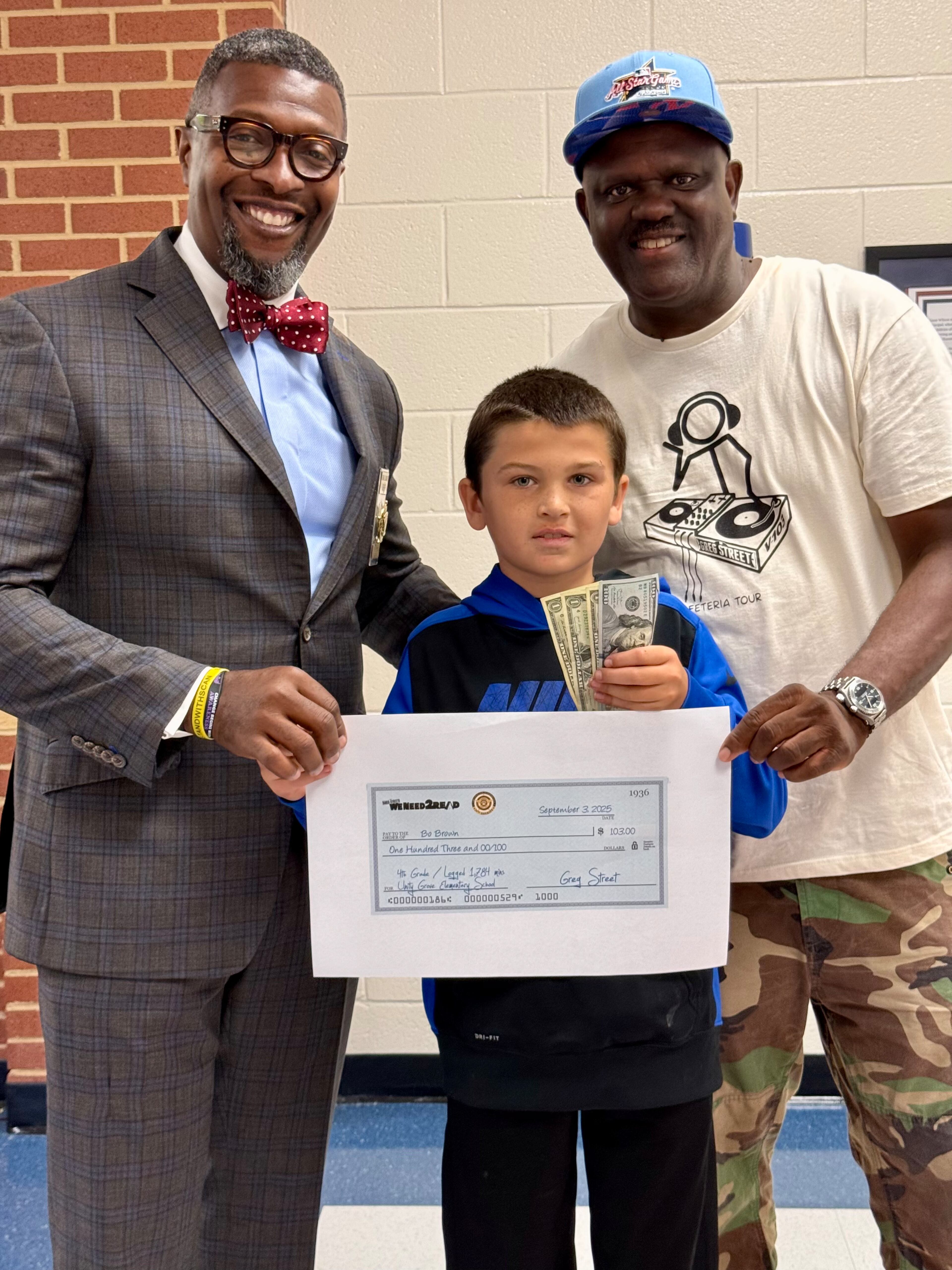 Greg Street (right) rewards fourth grade Bo Brown a WeNeed2Read reward of $103 at Unity Grove Elementary School in Locust Grove on Sept. 3, 2025. They are joined by Henry County sheriff Reginald Scandrett. (Courtesy)