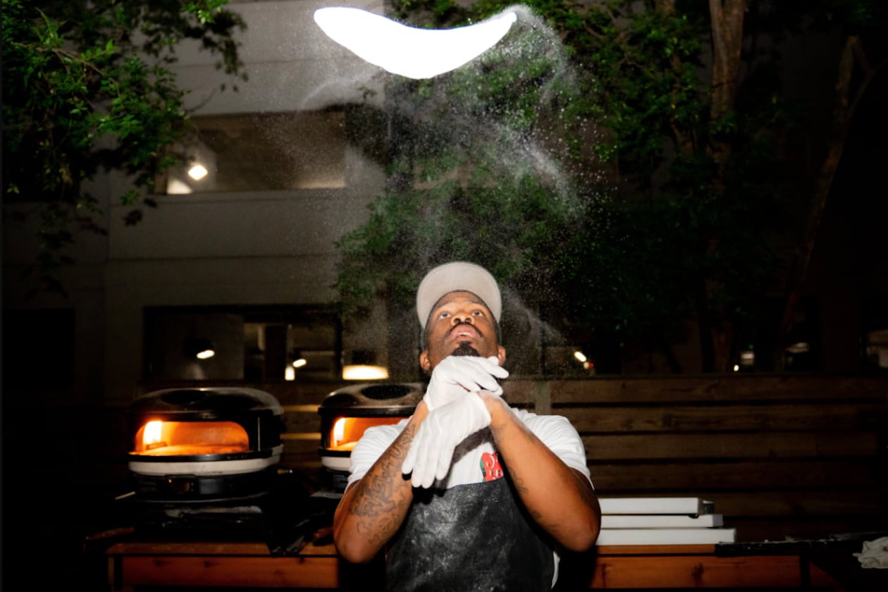 A dramatic photo showing Matthew Foster tossing pizza dough. The pizza dough spins high above Foster's head. Sprinkles of flour fly in the air between and around Foster and the dough.