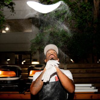 A dramatic photo showing Matthew Foster tossing pizza dough. The pizza dough spins high above Foster's head. Sprinkles of flour fly in the air between and around Foster and the dough.