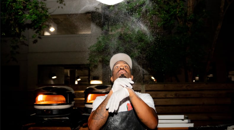 A dramatic photo showing Matthew Foster tossing pizza dough. The pizza dough spins high above Foster's head. Sprinkles of flour fly in the air between and around Foster and the dough.