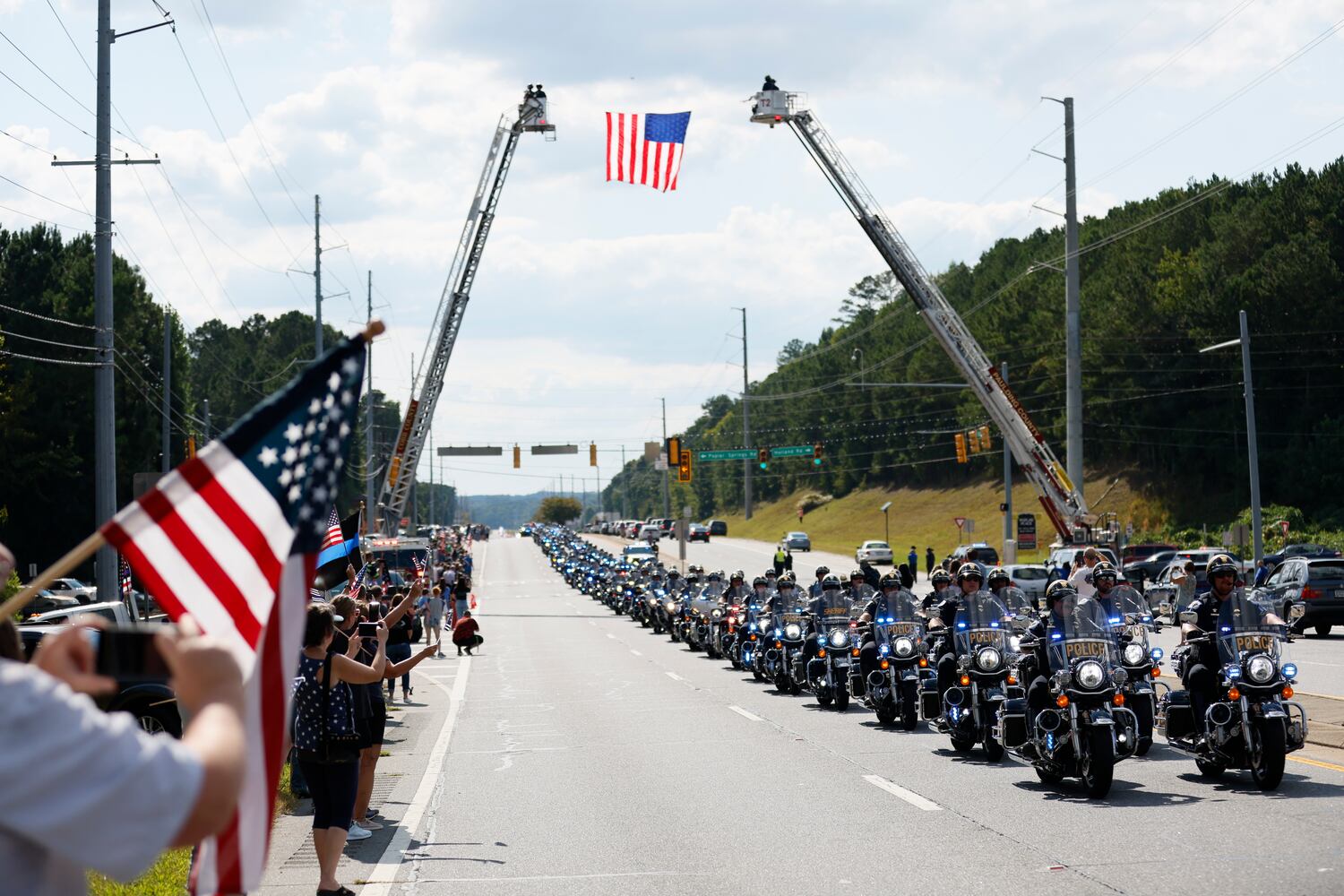 A motorcade leads the procession of fallen Marshall Samual Ervin Jr as people pay their respect with American flags over Dallas Highway on Thursday, September 15, 2022.  Marshall Samual Ervin Jr, alongside Cobb County Deputy Koleski, was killed late Thursday while attempting to arrest Christopher James Cook Jr. at home in the Hampton Glen subdivision last week. Miguel Martinez / miguel.martinezjimenez@ajc.com