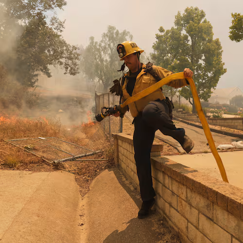A firefighter hops over a fence while battling the Sandy Fire Monday, May 18, 2026, in Simi Valley, Calif. (AP Photo/Ethan Swope)