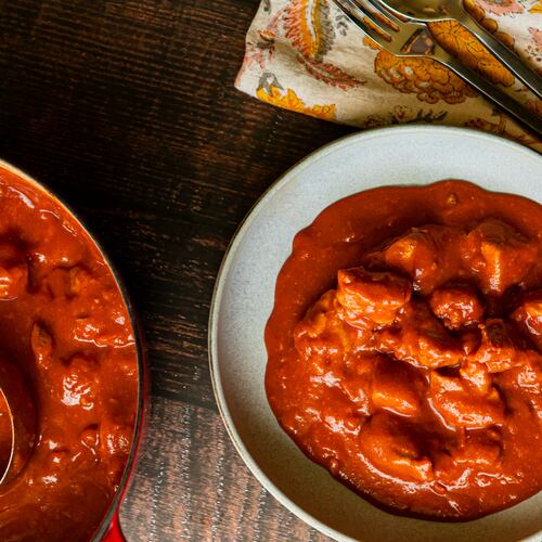 A bowl of butter chicken, also called murgh makhani, is in a bowl on a table. A skillet full of butter chicken is next to it, as is a cloth napkin with a fork and knife.