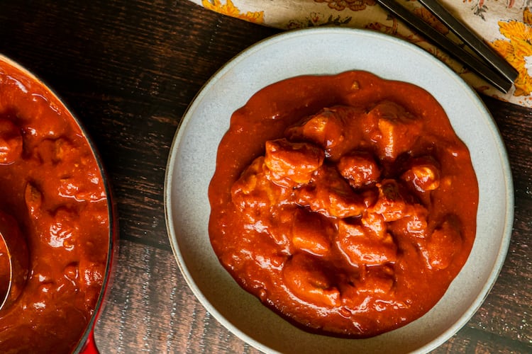 A bowl of butter chicken, also called murgh makhani, is in a bowl on a table. A skillet full of butter chicken is next to it, as is a cloth napkin with a fork and knife.