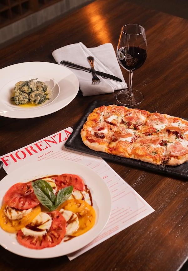 Several Italian items on a restaurant table, a caprese salad plate overlapping a menu with the name Fiorenza at the top, plus a glass of red wine to the right.