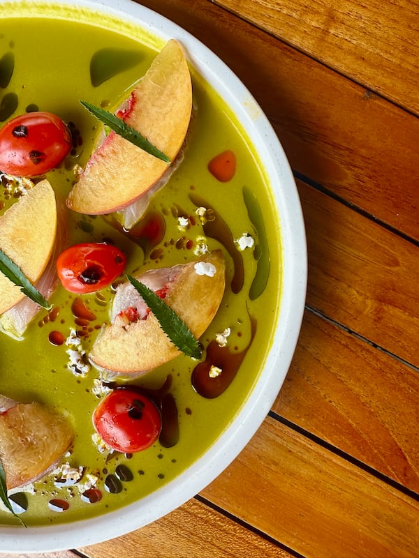 A close-up of a plate containing a colorful sauce dotted with peaches, hamachi and cherry tomatoes and sprinkled with herbs and oil.