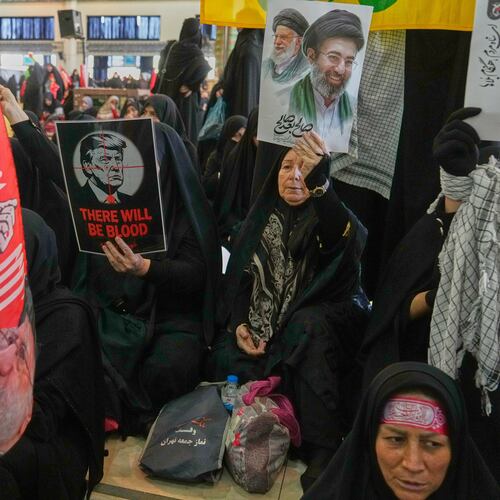 A woman holds a poster depicting U.S. President Donald Trump in the crosshairs of a rifle scope with the English words "There Will Be Blood," while another woman holds a portrait of the late Supreme Leader Ali Khamenei and his son, current Supreme Leader Mojtaba Khamenei, as mourners gather to commemorate the late leader at the Imam Khomeini Mosalla Grand Mosque in Tehran, Iran, Tuesday, July 14, 2026. (AP Photo/Vahid Salemi)