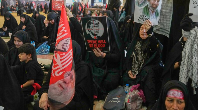 A woman holds a poster depicting U.S. President Donald Trump in the crosshairs of a rifle scope with the English words "There Will Be Blood," while another woman holds a portrait of the late Supreme Leader Ali Khamenei and his son, current Supreme Leader Mojtaba Khamenei, as mourners gather to commemorate the late leader at the Imam Khomeini Mosalla Grand Mosque in Tehran, Iran, Tuesday, July 14, 2026. (AP Photo/Vahid Salemi)