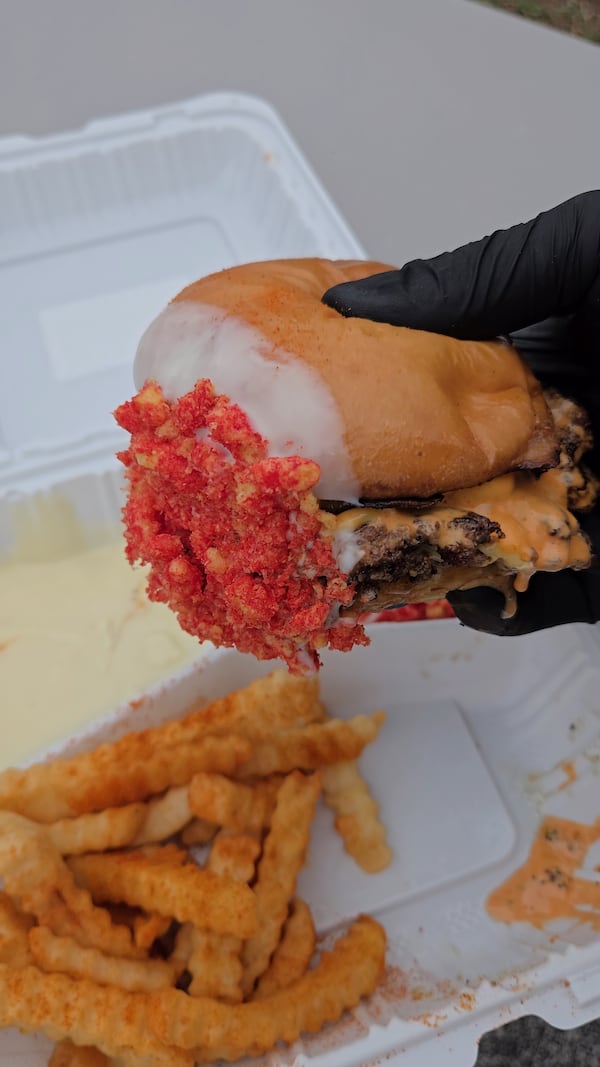 A hand holds a smash cheeseburger. The burger end has been dipped in cheese sauce and Flamin' Hot Cheetos crumbs, giving the burger a neon edge. French fries are shown below the burger.
