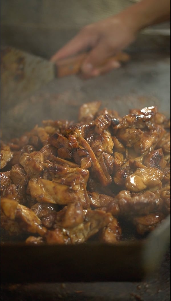 A close-up shot of pieces of glazed chicken on a smoking griddle, with a hand holding a spatula in the background.