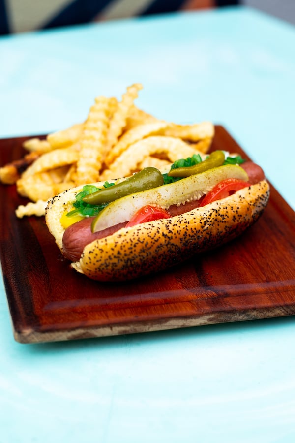 A hot dog on a sesame seed bun is loaded with a pickle spear, jalapeno peppers, yellow mustard and relish. French fries are next to the hot dog, both of which are on a wood-colored plate.