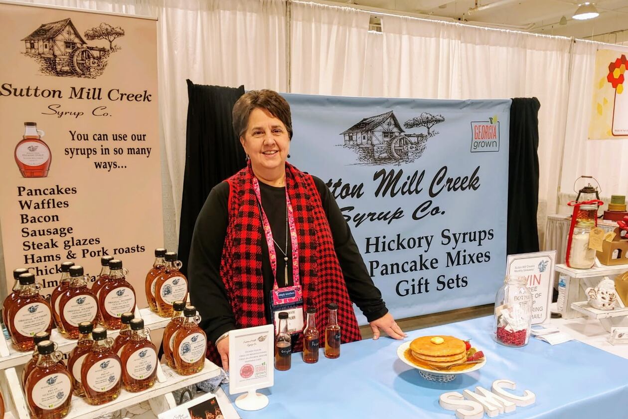 A woman in a red tartan scarf stands behind a table holding a display of hickory syrup with informational banners behind her from Sutton Mill Creek Syrup Co.