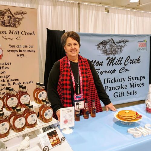 A woman in a red tartan scarf stands behind a table holding a display of hickory syrup with informational banners behind her from Sutton Mill Creek Syrup Co.