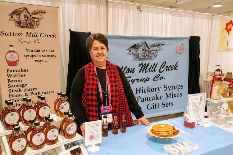 A woman in a red tartan scarf stands behind a table holding a display of hickory syrup with informational banners behind her from Sutton Mill Creek Syrup Co.