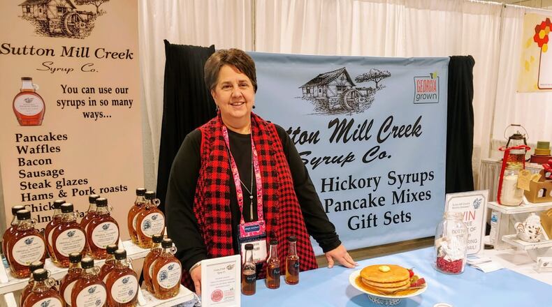 A woman in a red tartan scarf stands behind a table holding a display of hickory syrup with informational banners behind her from Sutton Mill Creek Syrup Co.