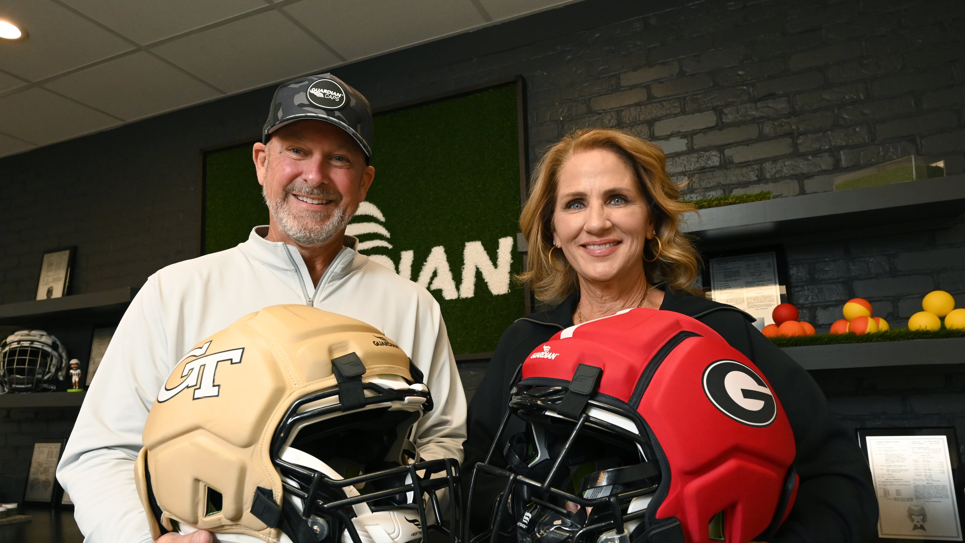Lee and Erin Hanson, co-founders of Guardian Sports, hold Guardian Caps with Georgia Tech and UGA logos as they pose for a photo at Guardian Sports' headquarters, Tuesday, Oct. 21, 2025, in Peachtree Corners. The Guardian Cap is the leading soft shell helmet cover engineered for impact reduction. (Hyosub Shin/AJC)