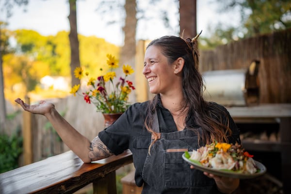 Ryanne Rothenberg smiles as she gestures to something off camera. She is standing outside, holding a colorful dish of vegetables and shrimp.