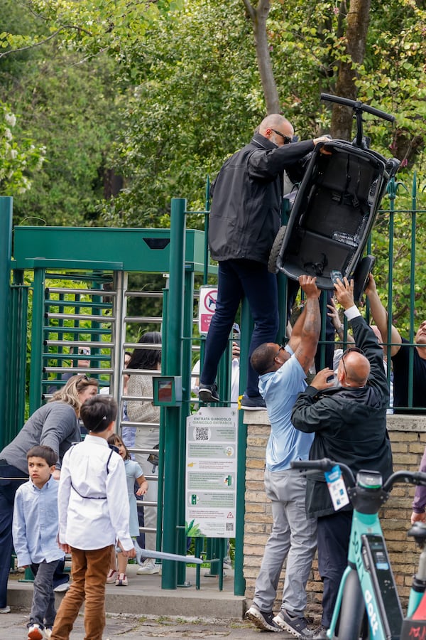 Security personnel accompanying U.S. Vice President JD Vance lift a trolley over the turnstiles at the exit of Rome's Botanic Gardens after a tour inside the gardens Vance did with his wife Usha, partially hidden behind the gate, their daughter Mirabel, sons Vivek, left, and Ewan, back to camera, Saturday, April 19, 2025. (AP Photo/Riccardo De Luca)