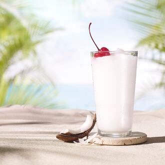 A tall, frozen pina colada in a stylized photo, with an island beach backdrop and a wedge of fresh coconut propped against the glass.