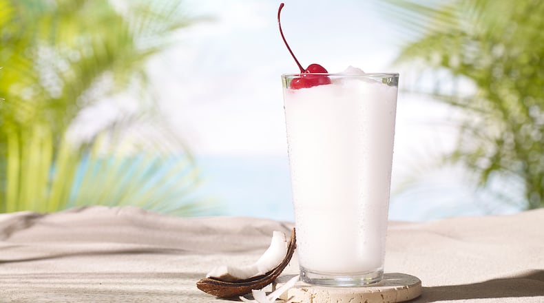 A tall, frozen pina colada in a stylized photo, with an island beach backdrop and a wedge of fresh coconut propped against the glass.